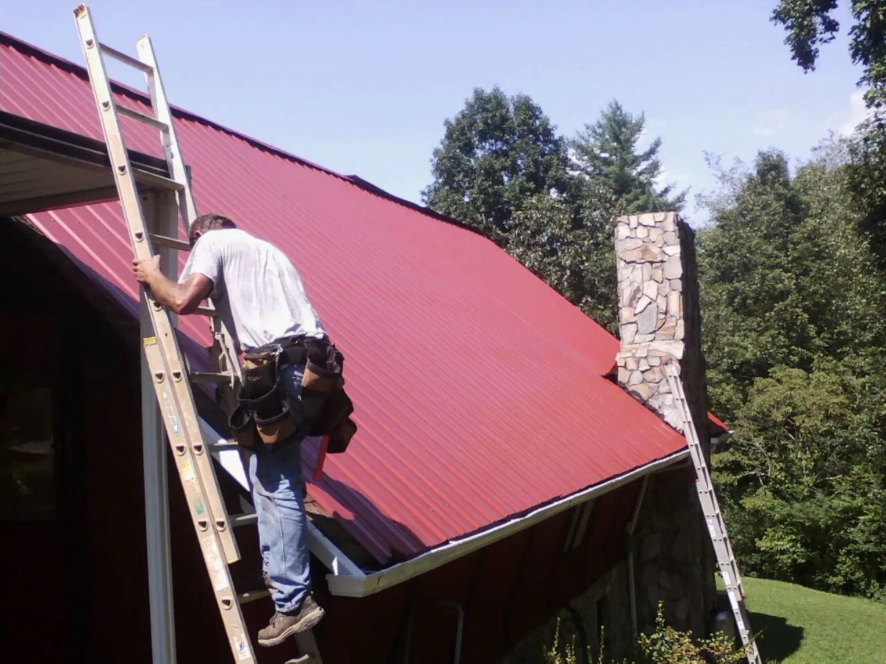 Roofer on a ladder beside a red metal roof and stone chimney — Soffit & Fascia Repair by Roofing Brothers in East Killingly